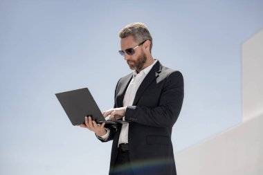 İş adamı dışarıda dizüstü bilgisayar kullanıyor. Dizüstü bilgisayarı olan olgun bir iş adamı. Dizüstü bilgisayarı olan ciddi bir iş adamı. İnternette iş. İş adamı laptoptaki verileri kontrol ediyor. Takım elbiseli iş adamı internette çalışıyor.