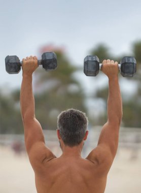 Spor adamının dambılları açık havada. Spor aktivitelerine katılan atletik bir adam. Formda adam geri döndü. Spor egzersizleri. Spor seansındaki üstsüz adam, geri döndü. Dambıllı açık hava eğitimi