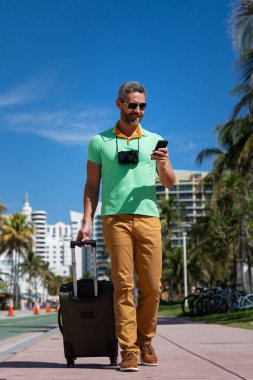 Miami sokaklarında bavulu ve kamerası olan bir turist. Bir erkek turist, tropikal bir şehirde yaz tatilinin tadını çıkarır. Yaz şehrinde bavullu bir turist. Erkek turist şehirde yürüyor