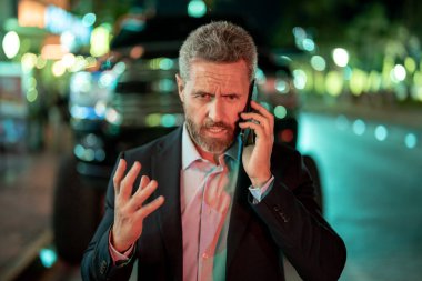 Kızgın iş adamı gece yarısı telefonda konuşuyor. Şehirde telefonu olan takım elbiseli üzgün bir adam. Telefon kullanan bir iş adamı. Şehir ışıkları yakınındaki bir iş adamından telefon geldi. Kötü iş çağrısı.