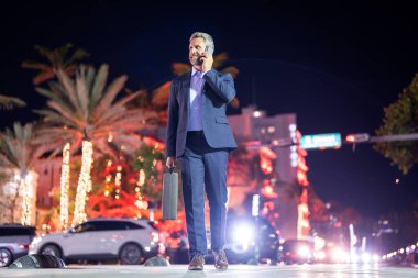Evrak çantası ve telefonda yürüyen bir iş adamı. Geceleri takım elbiseli zengin bir adam. Sokaktaki iş adamı. Gece şehrinin yakınlarında yürüyen bir iş adamı. İş stili. Lüks ortamda zengin bir işadamı
