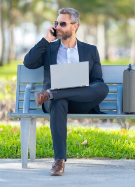 Kendine güvenen bir iş adamı bankta dizüstü bilgisayarı ve parktaki telefonuyla oturuyor. Başarılı bir iş adamı. Olgun iş adamı tatilin tadını çıkarıyor. İş adamı parktaki bankta mola veriyor. Yaz işleri.