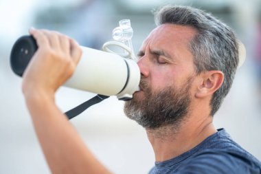Spor salonundaki adam bir bankta dinleniyor ve su şişesinden su içiyor. Antrenmandan sonra bir bankta oturan olgun bir spor modeli. Fitness sonrası ara veren bir adam spor su şişesinden su içer.