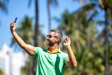 Mutlu turist palmiye ağacının yanında selfie çeker. Adam yaz tatilinde selfie çekiyor. Gezgin, tropikal bir yerde selfie çekiyor. Palmiye ağaçlı selfie tropikal titreşimleri yakalar.