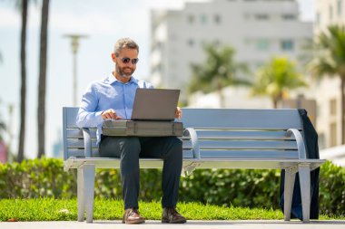 Kendine güvenen bir iş adamı bankta dizüstü bilgisayarı ve parktaki telefonuyla oturuyor. Zengin iş adamı. Olgun iş adamı tatilin tadını çıkarıyor. İş adamı parktaki bankta mola veriyor. Yaz işleri.