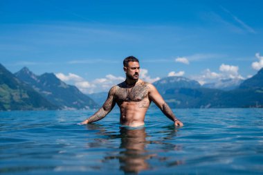 Seksi yaz erkeği. Suda ıslak kaslı erkek vücudu. Çıplak seksi erkek vücudu. İnsan yaz doğasında suya su sıçratır. Yaz plajında poz veren formda bir adam. Dağ gölünde su sıçratan güçlü bir adam. Yaz gezisi