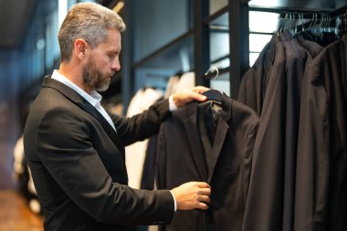 Takım elbiseli, butikte şık bir adam. Bir iş adamı mağazada şık bir takım elbise seçer. Modayı takip eden bir müşteri resmi kıyafet alırken ceketi inceler. Klasik kıyafetli çekici bir manken.