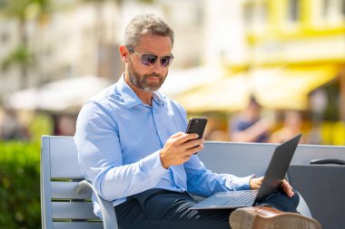 Kendine güvenen bir iş adamı bankta dizüstü bilgisayarı ve parktaki telefonuyla oturuyor. Zengin iş adamı. Olgun iş adamı tatilin tadını çıkarıyor. İş adamı parktaki bankta mola veriyor. Kentsel iş