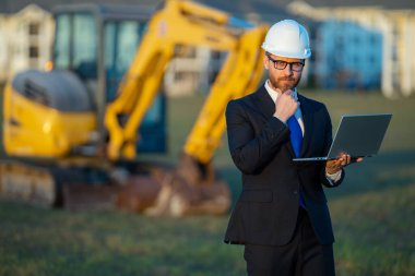 İnşaat mühendisi inşaat alanında çalışıyor. Olgun mühendis. İnşaat alanında takım elbiseli ve kasklı bir adam var. Orta yaşlı inşaat mühendisi kazıcının yanında duruyor.