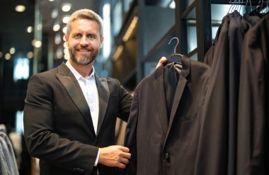 Olgun bir adam kıyafeti için mükemmel bir takım seçer. İyi giyimli bir erkek model bir butikte poz veriyor. Perakende satış asistanı şık bir adamın mükemmel takımı bulmasına yardım eder. Mağaza müdürü şık bir takım elbise ayarlar.