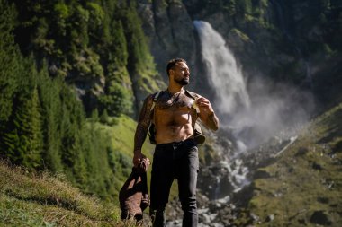 Yaz tatilindeki erkek turist. Yaz tatilinde çıplak gövdeli yakışıklı bir adam. Dışarıda seksi bir yürüyüşçü var. Şelale manzarasında yürüyen bir turist. İnsan Seyahati ve doğa