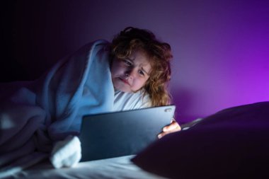 Tabletteki çocuk yatağı hikâyesi. Çocuk yatakta uzanıyor ve karanlık odada tablet üzerinde internette sörf yapıyor. Geceleri Tablet okuyan bir çocuk. E-kitap, hikaye ve çocuk uygulaması oyunu. Yatmadan önce tablet kullanan bir çocuk.