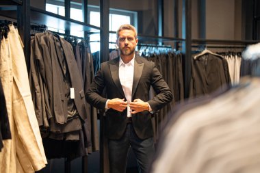 Bir adam butikte alışveriş yaparken klasik bir terzi kıyafeti giyer. Başarılı bir iş adamı için klasik bir kıyafet. Takım elbiseli modaya uygun bir mağaza. Takım elbise seçen bir iş adamı. Bir erkek resmi etkinlik için takım elbise seçer.