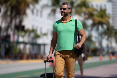 Tropikal şehirde bavulları olan bir gezgin. Bir erkek gezgin, tropik bir şehirde yaz tatilinin tadını çıkarır. Yaz şehrinde bavullu bir turist. İnsan gezgin şehirde yürür