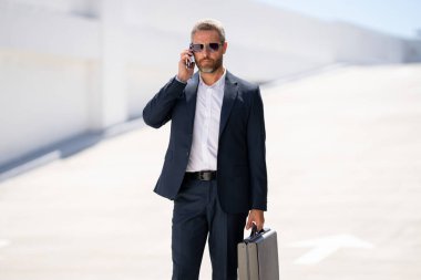 İş portresi. Dışarıdaki telefondan iş adamı aradı. Başarılı lider hareket halinde. Takım elbiseli, çantalı bir adam telefonda konuşuyor. İşadamı işe gidiyor. Çantası olan bir iş adamı. İş başarısı
