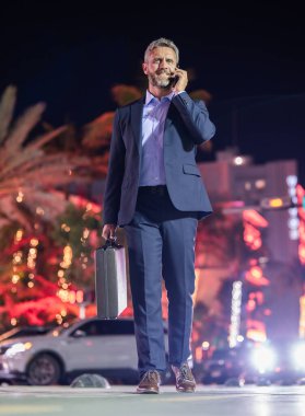 Evrak çantası ve telefonda yürüyen bir iş adamı. Geceleri takım elbiseli zengin bir adam. Sokaktaki iş adamı. Gece şehrinin yakınlarında yürüyen bir iş adamı. İş stili. Lüks ortamda zengin bir işadamı