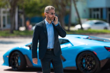 Dışarıdaki iş adamı. Zengin iş adamı başarının tadını çıkarıyor. Şık takım elbiseli lüks iş adamı. Pahalı arabanın önündeki zengin adam. Lüks iş seyahati. Klasik takım elbiseli, kendinden emin zengin bir iş adamı.