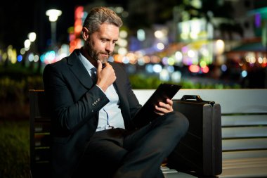 Takım elbiseli bir adam dışarıda tablet üzerinde çalışıyor. Tabletli bir iş adamı. Gece sokağında tabletli bir iş adamı. Tablet kullanan bir işadamı, bankta oturur. Gece işi.