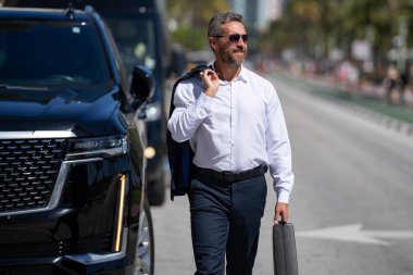 Lüks bir arabanın yanında zengin bir iş adamı. Takım elbiseli zengin bir iş adamı. Şehirdeki başarılı zengin iş adamı. Kendine güvenen bir iş adamı. Başarı işi. İş adamı sokakta yürüyor.