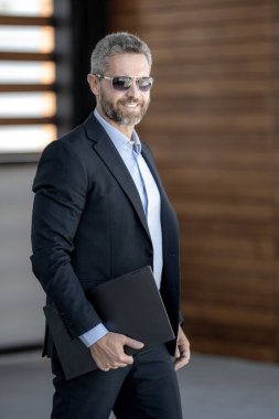 Başarılı bir iş adamı dışarıda dizüstü bilgisayarla dolaşır. Kendine güvenen iş adamı dizüstü bilgisayarı dışarıda tutuyor. Takım elbiseli, olgun, serbest çalışan bir iş adamı. Başarı işi. Takım elbiseli zengin iş adamı