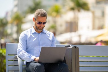 Kendine güvenen işadamı bankta dizüstü bilgisayarı ve parktaki telefonuyla oturuyor. Zengin iş adamı. Olgun iş adamı tatilin tadını çıkarıyor. İş adamı parktaki bankta mola veriyor. Kentsel iş