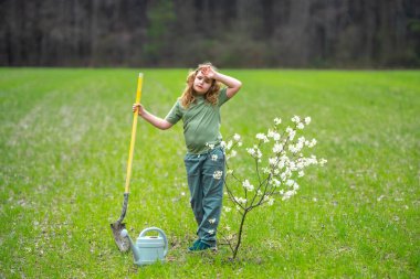 Çocuk dışarıda ağaç dikiyor. Bitkilerin bahar etkinliği. Çocuk kürek ve su bidonuyla çalışıyor. Çocuk meyve ağacı dikmiş. Bahçıvan çocuklar. Çocuk yetiştirme bitkisi ve ağaç bakımı