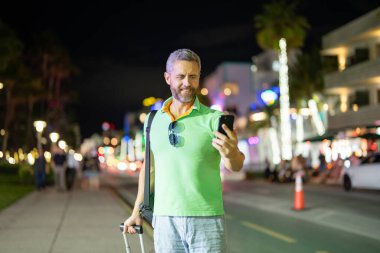 Gece Miami sokaklarında telefon kullanan bir gezgin. Erkek turist yaz macerası sırasında telefonla arıyor. Telefonda sıradan bir turist konuşması. Gezgin telefonunu kontrol ediyor. Tropik bir şehirde bavullu bir adam.