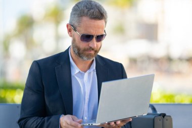 Kendine güvenen işadamı bankta dizüstü bilgisayarı ve parktaki telefonuyla oturuyor. Zengin iş adamı. Olgun iş adamı tatilin tadını çıkarıyor. İş adamı parktaki bankta mola veriyor. Kentsel iş