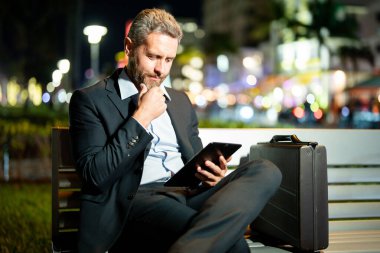 Takım elbiseli bir adam dışarıda tablet üzerinde çalışıyor. Tabletli bir iş adamı. Gece sokağında tabletli bir iş adamı. Tablet kullanan bir işadamı, bankta oturur. Gece işi.