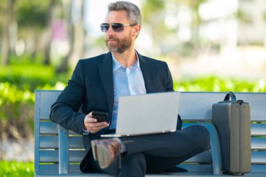 Kendine güvenen bir iş adamı bankta dizüstü bilgisayarı ve parktaki telefonuyla oturuyor. İspanyol iş adamı. Olgun iş adamı tatilin tadını çıkarıyor. İş adamı parktaki bankta mola veriyor. Yaz işleri.