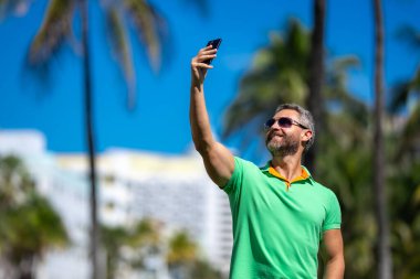 Turist palmiye ağacının yanında selfie çeker. Adam yaz tatilinde selfie çekiyor. Gezgin, tropikal bir yerde selfie çekiyor. Palmiye ağaçlı selfie tropikal titreşimleri yakalar.
