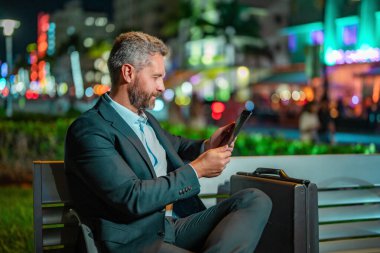 Çevrimiçi gece işi. Dışarıda tablet üzerinde çalışan bir iş adamı. Tabletli bir iş adamı. Gece sokağında tabletli bir iş adamı. Tablet kullanan bir işadamı, bankta oturur. Gece işi.