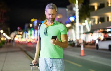 Gece Miami sokaklarında telefon kullanan bir gezgin. Erkek turist yaz macerası sırasında telefonla arıyor. Telefonda sıradan bir turist konuşması. Gezgin telefonunu kontrol ediyor. Tropik bir şehirde bavullu bir adam.