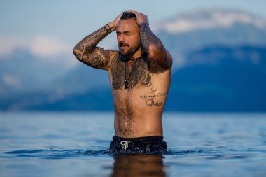 Seksi yaz erkeği. Suda ıslak kaslı erkek vücudu. Çıplak seksi erkek vücudu. İnsan yaz doğasında suya su sıçratır. Yaz plajında poz veren formda bir adam. Dağdaki gölde su sıçratan güçlü adam.