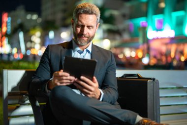 İnternet işi. Dışarıda tablet üzerinde çalışan bir iş adamı. Tabletli bir iş adamı. Gece sokağında tabletli bir iş adamı. Tablet kullanan bir işadamı, bankta oturur. Gece işi. Uzak çalışma