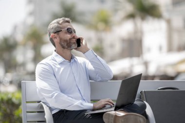 Kendine güvenen bir iş adamı bankta dizüstü bilgisayarı ve parktaki telefonuyla oturuyor. İspanyol iş adamı. Olgun iş adamı tatilin tadını çıkarıyor. İş adamı parktaki bankta mola veriyor. Yaz işleri.