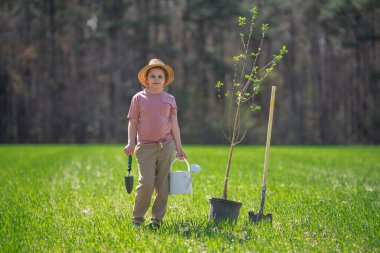 Kürekle ağaç yetiştiren çocuk. Arka bahçede ağaç yetiştiren çocuk. Bahar tarlasında ağaç yetiştiren çocuk. Sulama kutusu ve küreği olan küçük bahçıvan.