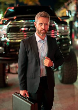 Gece vakti evrak çantalı bir iş adamı. Şehirde takım elbiseli zengin bir adam. Lüks arabanın yanında bir iş adamı. Şehir ışıklarının yanında yürüyen bir iş adamı. İş stili. Lüks ortamda zengin bir işadamı