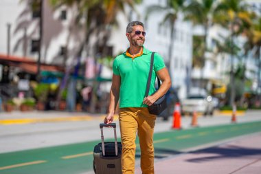 Tropikal şehirde bavul taşıyan bir turist. Yaz tatilinde bir erkek turist. Yaz şehrinde bavullu bir turist. Erkek turist şehirde yürüyor. Seyahat yaşam tarzı. Yaz tatili kaçamağı