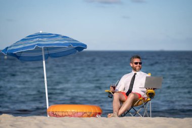 Yaz tatillerinde başarılı bir iş adamı. Komik iş adamı deniz sahilinde yaz kokteyli içip dizüstü bilgisayar kullanıyor. Yaz işi konsepti. Sıcak yaz. Çılgınca. Uzak çevrimiçi çalışma
