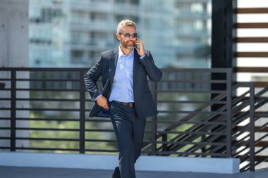 İş adamı telefonda konuşuyor ve yürüyor. Kendine güvenen bir iş adamı telefonla dışarı çıkar. İş telefonu konuşması. Şirket lideri telefonda konuşmakla meşgul. Zengin iş adamı. İş çağrısı