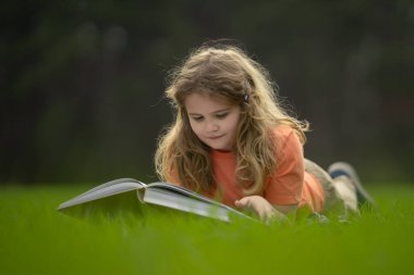 Çimlerin üzerinde kitap okuyan bir çocuk. Çocuk açık havada kitap okumaktan zevk alıyor. Güneşli bir günde çayırda kitapla ders çalışan bir çocuk. Doğada öğrenme. Okul çocuğu parkta kitap okuyor.