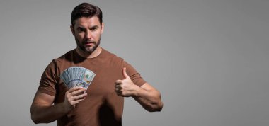 İyi ödeme. Yakışıklı adam gri stüdyoda izole edilmiş para tutuyor. Dolar banknotları halinde para. Yığınla para, finans, yatırım ve tasarruf. Model pankartı