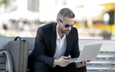 Kendine güvenen bir iş adamı bankta dizüstü bilgisayarı ve parktaki telefonuyla oturuyor. Başarılı bir iş adamı. Olgun iş adamı tatilin tadını çıkarıyor. İş adamı parktaki bankta mola veriyor. Yaz işleri.