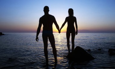 Gün batımında deniz kenarında bir çift silueti. Sevgililerle romantik plaj günbatımı. Sahilde birbirine aşık bir çift. Kumsalın yakınında bir çift romantik. Çift denize gidiyor. Birbirine aşık çiftler için yaz denizi