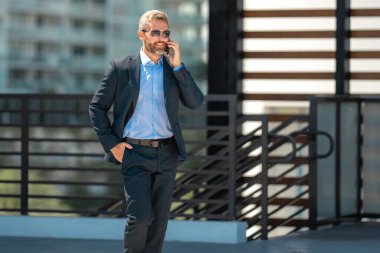 İş adamı telefonda konuşuyor ve yürüyor. Kendine güvenen bir iş adamı telefonla dışarı çıkar. İş telefonu konuşması. Şirket lideri telefonda konuşmakla meşgul. Zengin iş adamı. İş çağrısı