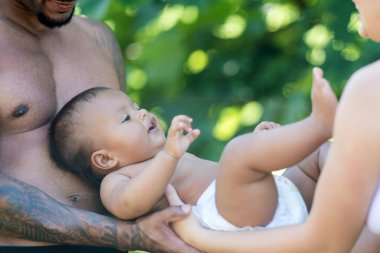 Genç çoklu etnik aile açık hava portresi. Karışık ırk ebeveynleri ve bebek birlikte vakit geçiriyorlar kucaklaşıyorlar ve parkta öpüşüyorlar. Afrika kökenli Amerikalı koca ve Kafkasyalı eş ve küçük çocuk yaz gününün tadını çıkarıyorlar.