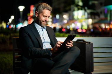 Dışarıda tablet üzerinde çalışan bir iş adamı. Tabletli bir iş adamı. Gece sokağında tabletli bir iş adamı. Tablet kullanan bir işadamı, bankta oturur. Gece işi.