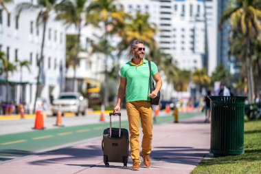 Tropikal şehirde bavulları olan bir gezgin. Bir erkek gezgin, tropik bir şehirde yaz tatilinin tadını çıkarır. Yaz şehrinde bavullu bir turist. İnsan gezgin şehirde yürür