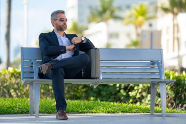 Kendine güvenen işadamı bankta dizüstü bilgisayarı ve parktaki telefonuyla oturuyor. Zengin iş adamı. Olgun iş adamı tatilin tadını çıkarıyor. İş adamı parktaki bankta mola veriyor. Kentsel iş